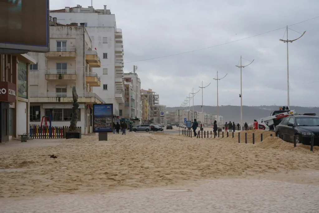Vue de l'avenue de la plage à Nazaré, au Portugal, après une forte tempête. La rue et le trottoir sont entièrement recouverts de sable, avec quelques piétons marchant au milieu de la voie ensablée. Au premier plan à droite, une voiture noire est partiellement ensablée et derrière elle, un engin de chantier (type Bobcat) travaille à déblayer le sable.