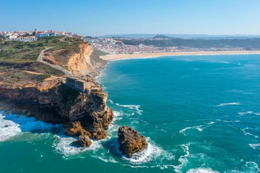 Vue aérienne spectaculaire de Nazaré au Portugal, montrant les falaises du Sitio avec le fort, la grande plage de sable, la ville et le port de pêche au loin sous un ciel bleu.