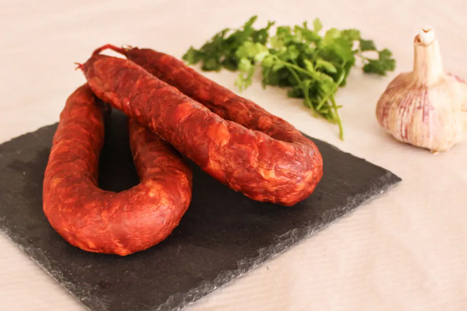 Présentation élégante d'un Chouriço portugais artisanal piquant 220 g de Quintinha d'Aldeia, posé sur une plaque d'ardoise noire avec une tête d'ail et des feuilles de persil frais en garniture.