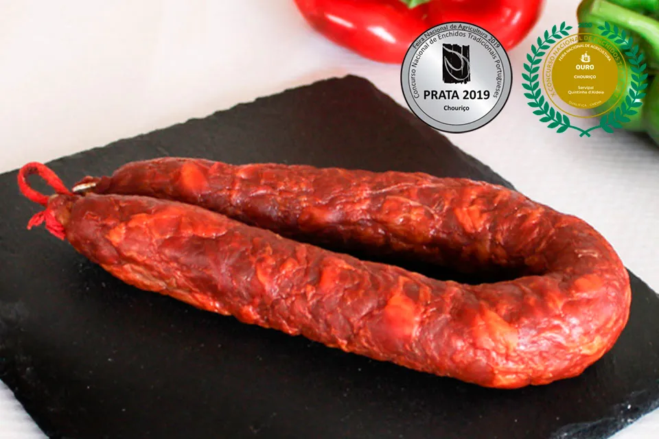 Chouriço portugais artisanal traditionnel Extra 220 g de Quintinha d'Aldeia présenté entier en forme de fer à cheval sur une ardoise noire, avec poivrons en arrière-plan et ses médailles aux concours nationaux : médaille d'argent 2019 et médailles d'or 2020 et 2024.