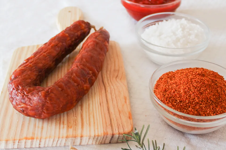 Chouriço portugais artisanal piquant 220 g de Quintinha d'Aldeia présenté sur une planche en bois, entouré de récipients contenant du paprika, du gros sel et de la pâte de piment pour illustrer ses ingrédients traditionnels.