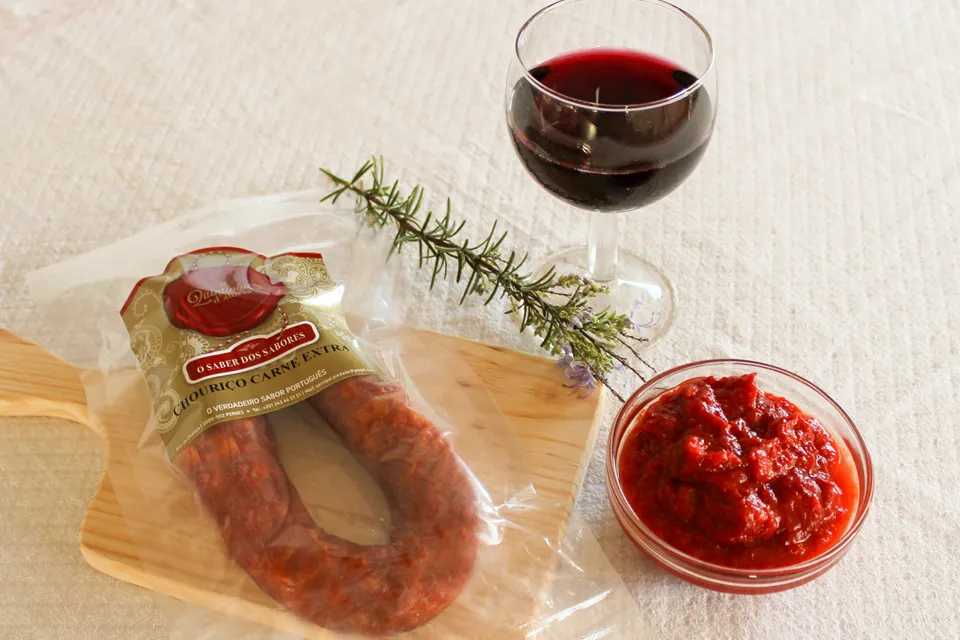Mise en scène de dégustation du Chouriço portugais artisanal traditionnel Extra 220 g de Quintinha d'Aldeia emballé sous vide sur une planche en bois, accompagné d'un verre de vin rouge, d'un ramequin de pâte de poivron (massa de pimentão) et d'un brin de romarin.