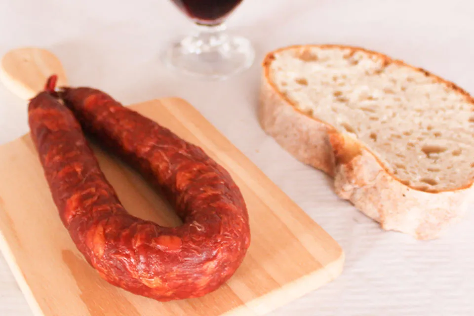 Suggestion de dégustation d'un Chouriço portugais artisanal piquant 220 g de Quintinha d'Aldeia sur planche en bois, accompagné d'une tranche de pain traditionnel et d'un verre de vin rouge en arrière-plan.