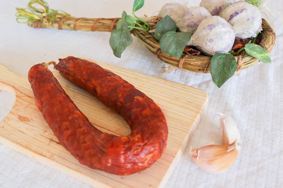 Présentation culinaire du Chouriço portugais artisanal traditionnel Extra 220 g de Quintinha d'Aldeia, entier et disposé sur une planche à découper en bois clair, accompagné de gousses d'ail frais et d'une tresse d'ail décorative en arrière-plan.