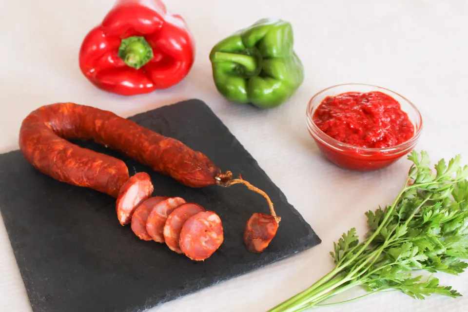 Chouriço portugais artisanal piquant 220 g de Quintinha d'Aldeia partiellement tranché sur une ardoise, entouré de poivrons rouge et vert, d'un ramequin de pâte de piment et de persil frais.