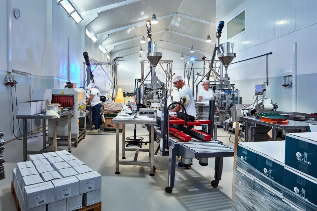 Vue d'ensemble de l'usine de conditionnement et d'emballage des produits Necton au Portugal. Des opérateurs en tenue de travail s'activent autour des lignes d'emballage modernes, des ensacheuses et des convoyeurs, au milieu de cartons Necton prêts à l'expédition.