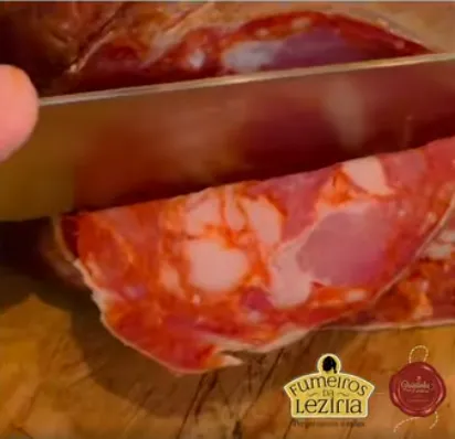 Gros plan sur la découpe d'une tranche de Paiola, gros saucisson sec fumé portugais artisanal de Quintinha d'Aldeia, par un couteau sur une planche en bois.